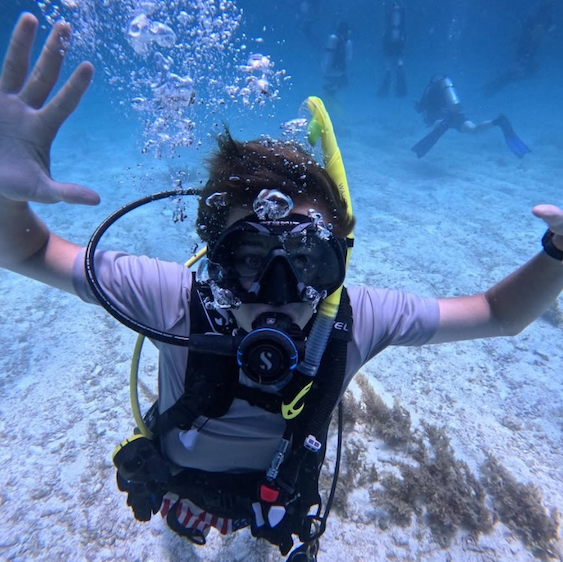 A scout scuba diving underwater over a sandy sea floor, arms outstretched as other divers explore behind